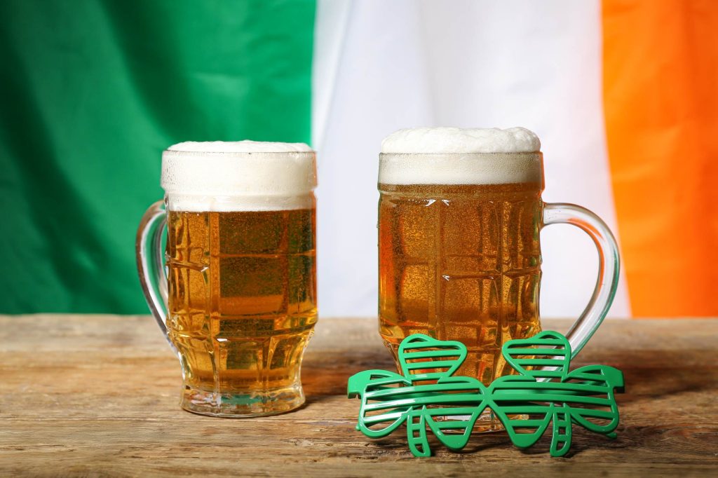 Mugs of beer and plastic eyeglasses on wooden table against flag of Ireland. St. Patrick's Day celebration